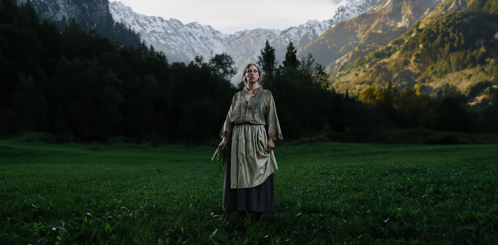 The woman standing in a meadow below snow-covered peaks