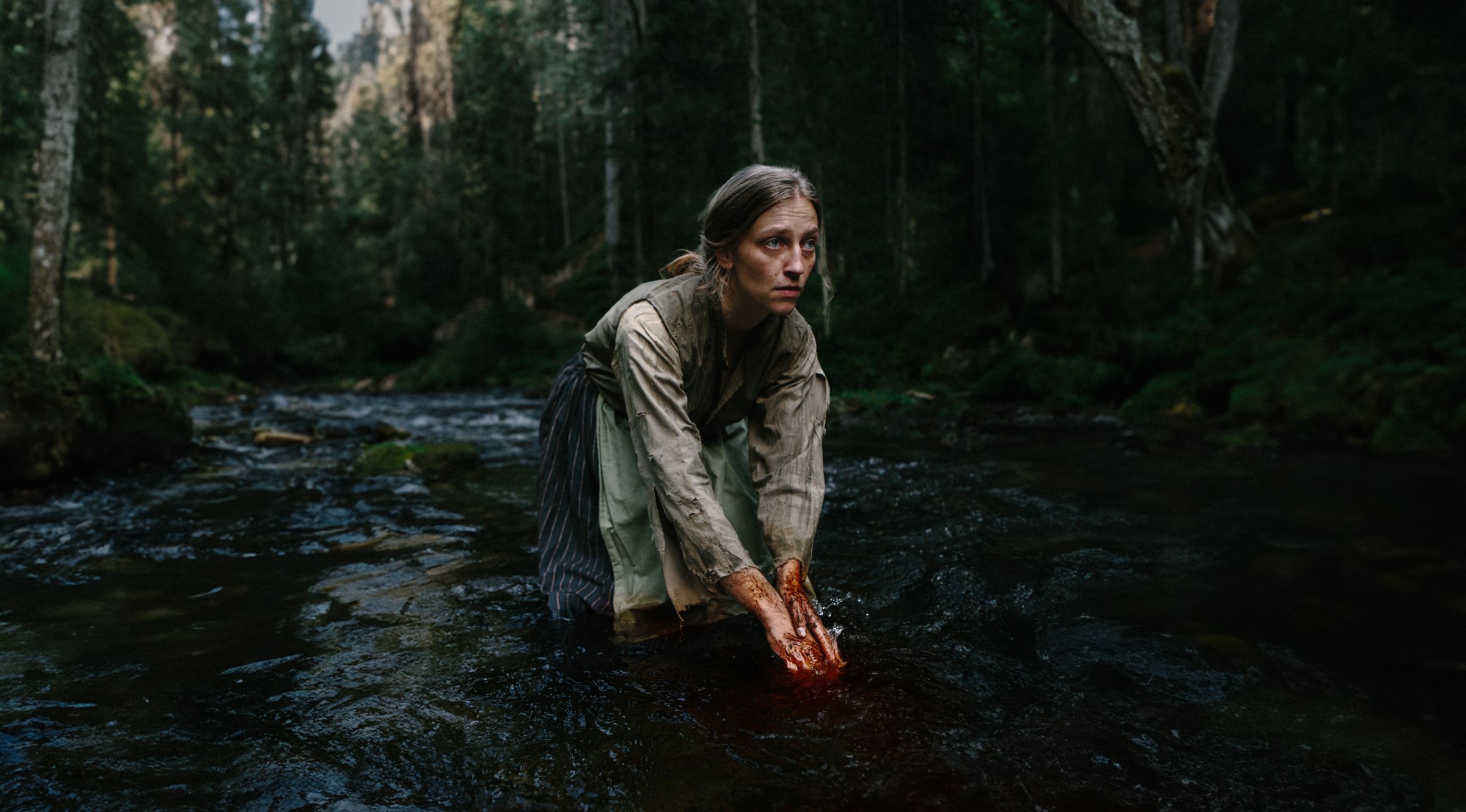 A woman kneeling in a mountain river, washing the blood from her hands